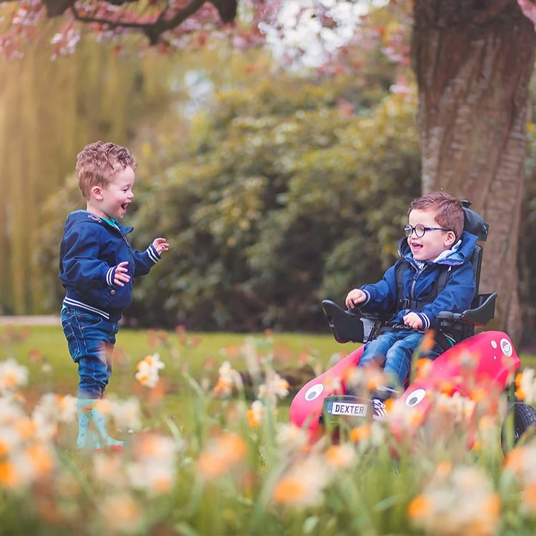 To små gutter som leker ute blant blomster; den ene står og ler, mens den andre sitter i en rød rullestol og smiler.