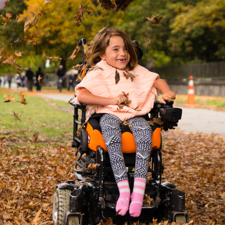 Jente i rullestol som leker blant høstløv – bevegelsesglede og frihet utendørs.