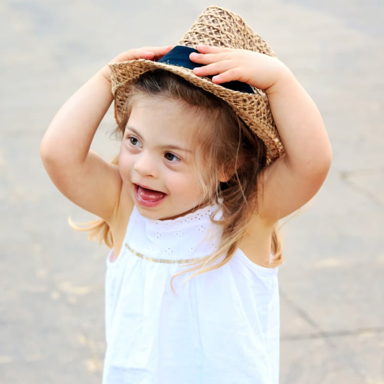 Lita jente med Downs syndrom smiler og holder en stråhatt på hodet mens hun leker utendørs.