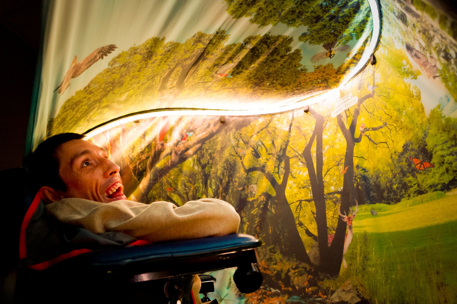 Bruker i rullestol smiler under Snoezle-telt med dagslys-effekt og trær på duken.