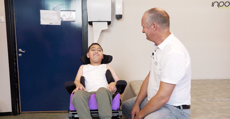 Et ungt barn sitter i en tilpasset rullestol og smiler til en mann som står ved siden av, sannsynligvis i en hyggelig samtale. Begge er innendørs, med mannen iført en hvit poloskjorte med 'inpo'-logoen, noe som antyder en terapeutisk eller klinisk setting