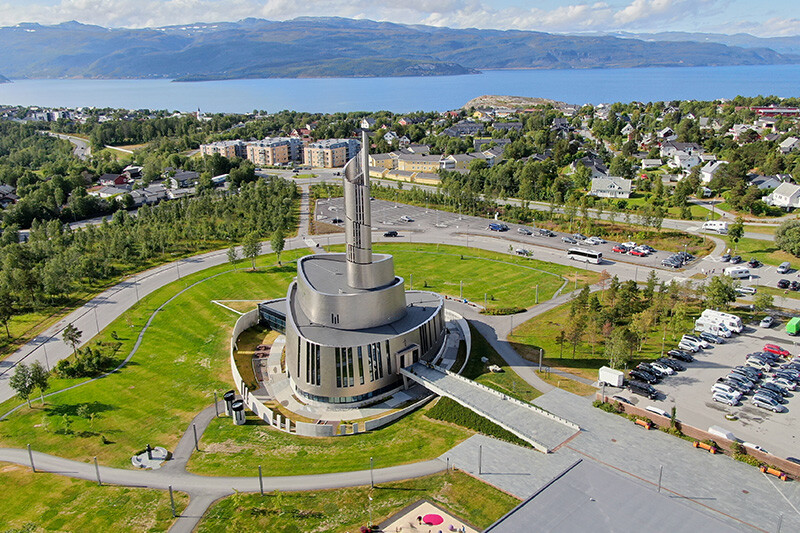 Nordlyskatedralen i Alta, en moderne kirke med spiralformet tårn, omgitt av grønt landskap og utsikt mot fjorden.