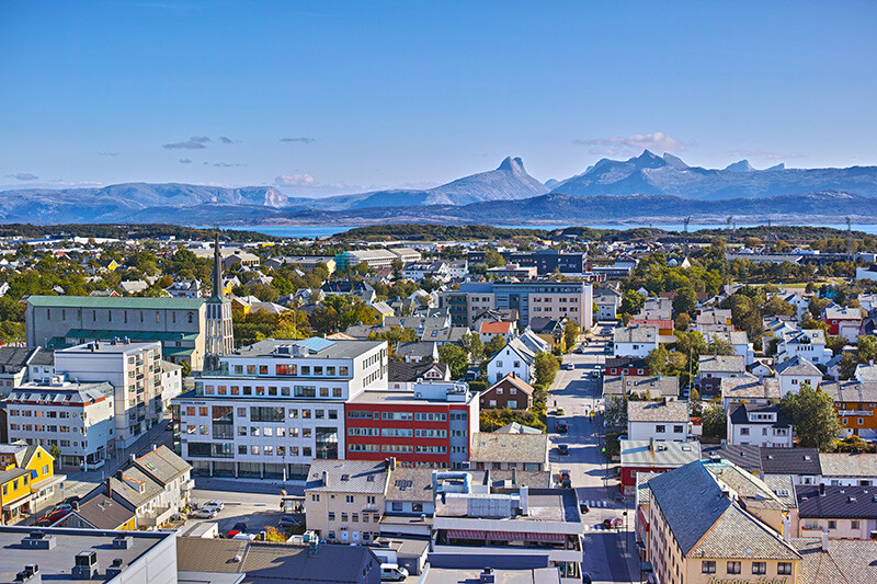 Panoramautsikt over Bodø med moderne og tradisjonelle bygninger, en kirke og majestetiske fjell i bakgrunnen.