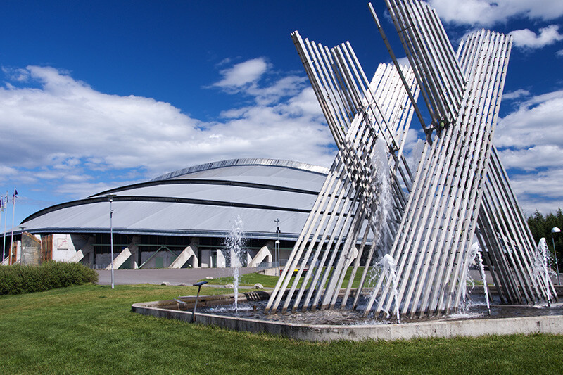 Vikingskipet i Hamar, en ikonisk skøytehall med en moderne fonteneskulptur i forgrunnen under en blå himmel.