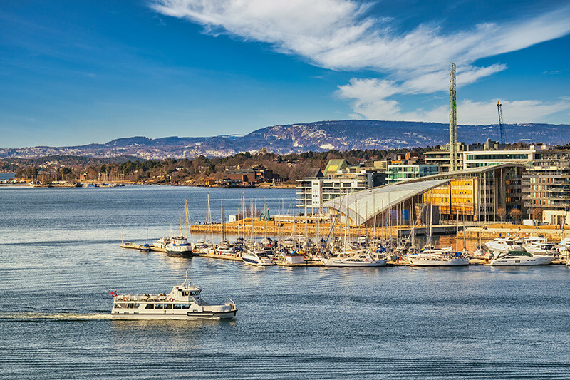 Oslofjord med båter og ferge foran Astrup Fearnley Museet for Moderne Kunst, med åser og blå himmel i bakgrunnen.