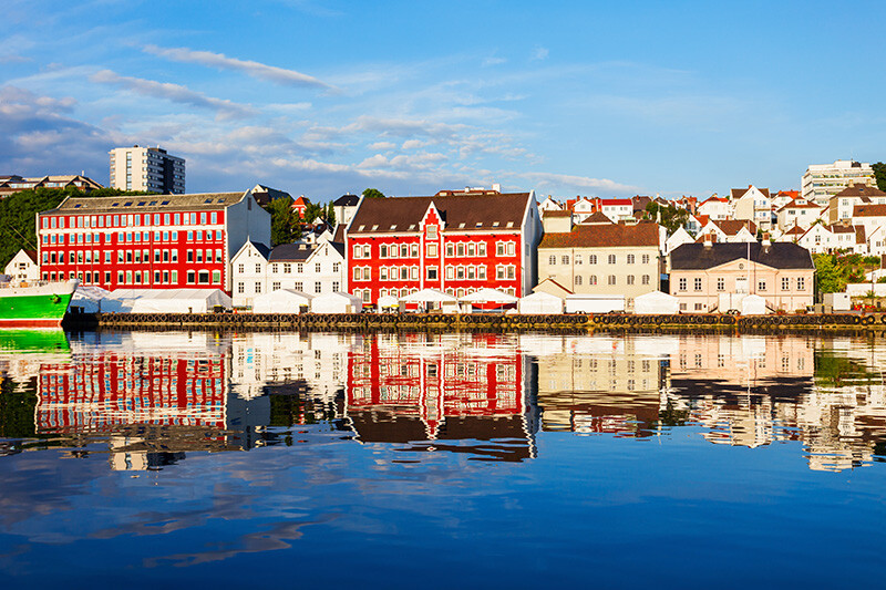 Fargerike trehus langs en havnefront speiler seg i det rolige vannet under en blå himmel i Stavanger.
