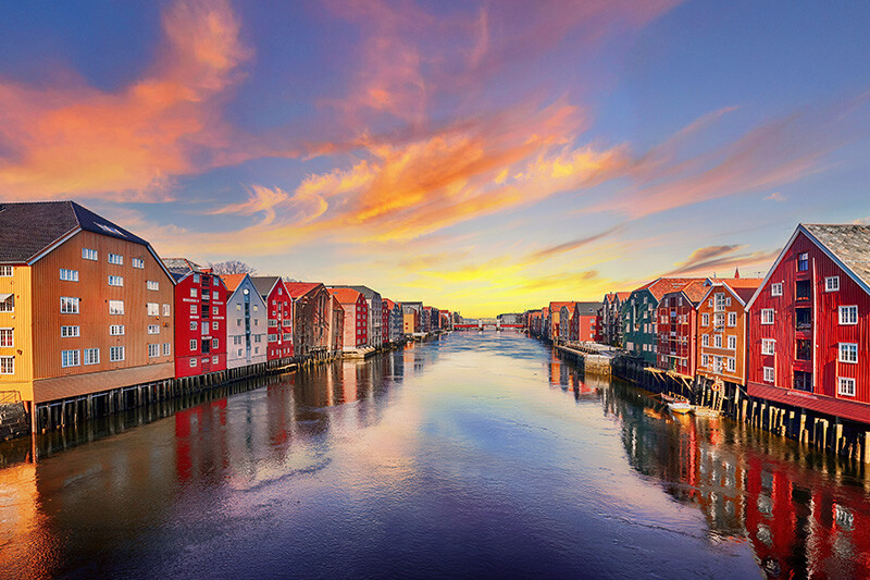 Bryggene langs Nidelva i Trondheim ved solnedgang, med fargerike trehus speilet i vannet.
